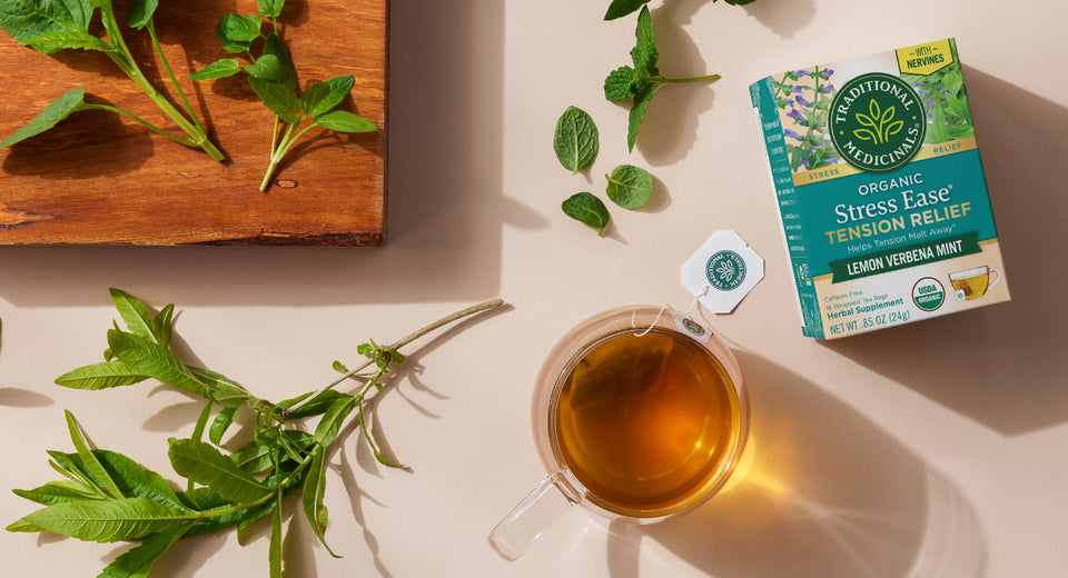 Cup of brewed tea and box of Stress Ease Tension Relief on table with herbs