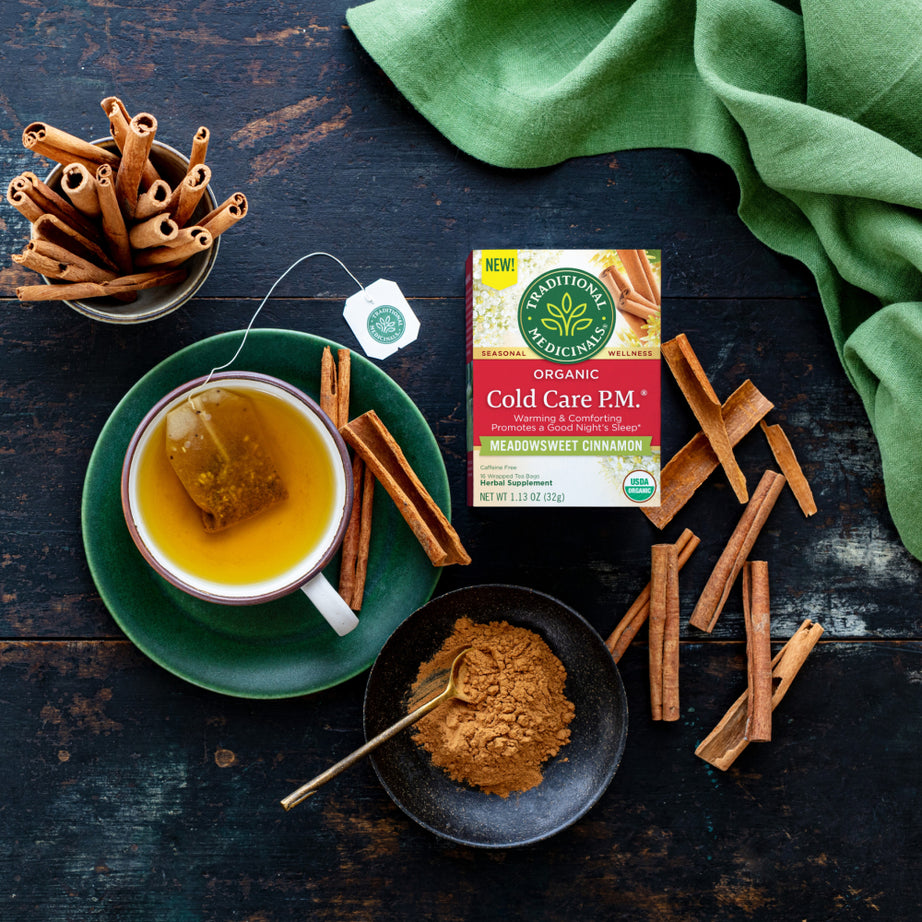 A cup of tea with cinnamon sticks and powder is placed on a dark table, next to a box of Cold Care P.M.® Tea, an organic nighttime blend that promotes a good night's sleep.