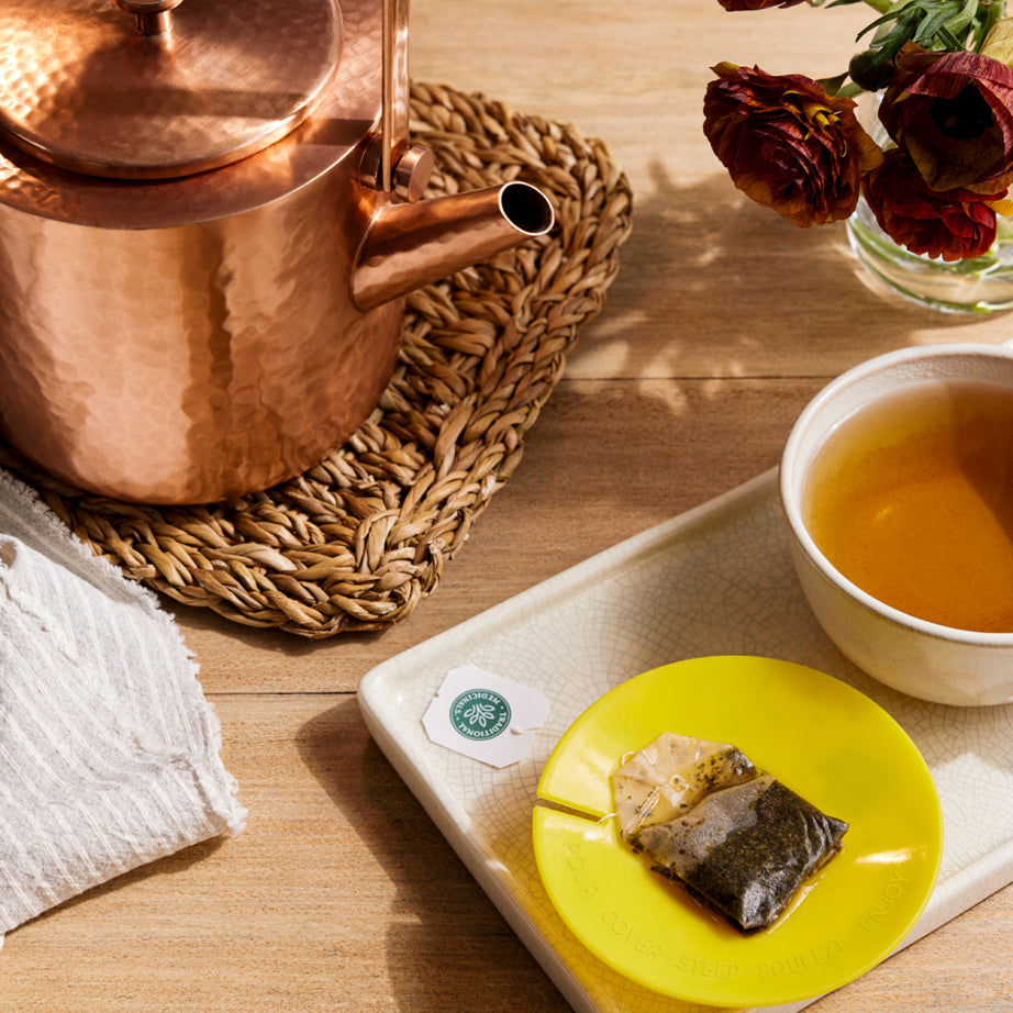 Copper kettle, Dandelion Leaf & Root Tea bag on a yellow dish, and a cup of tea promoting kidney function placed on a tray with flowers atop a wooden table.