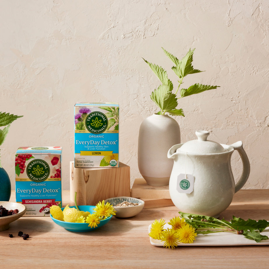 A tea setup featuring EveryDay Detox® Lemon Tea boxes from Traditional Medicinals, accompanied by a teapot, a vase with yellow flowers, and lemons, is arranged on a wooden table. It's an ideal choice for individuals looking to support liver and kidney function while promoting vibrant health and healthy skin.