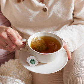 A person wearing a white shirt sits comfortably on a cozy blanket, savoring a soothing cup of Ginger & Chamomile Tea with a tea bag.