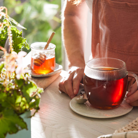 A person holding a steaming cup of Ginger & Chamomile Tea with a jar of honey nearby on a sunny table surrounded by plants, perfect for soothing indigestion.