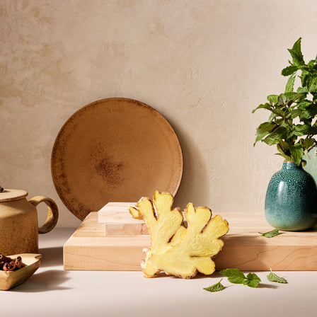 A vase filled with mint and organic ginger root accompanies rustic pottery on a wooden surface, creating a tranquil scene that subtly suggests the comforting benefits of Ginger Tea for promoting healthy digestion.