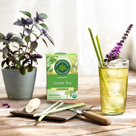 A box of Green Tea Lemongrass sits beside iced tea, lemongrass stalks, herbs, and a plant on a sunny white table.