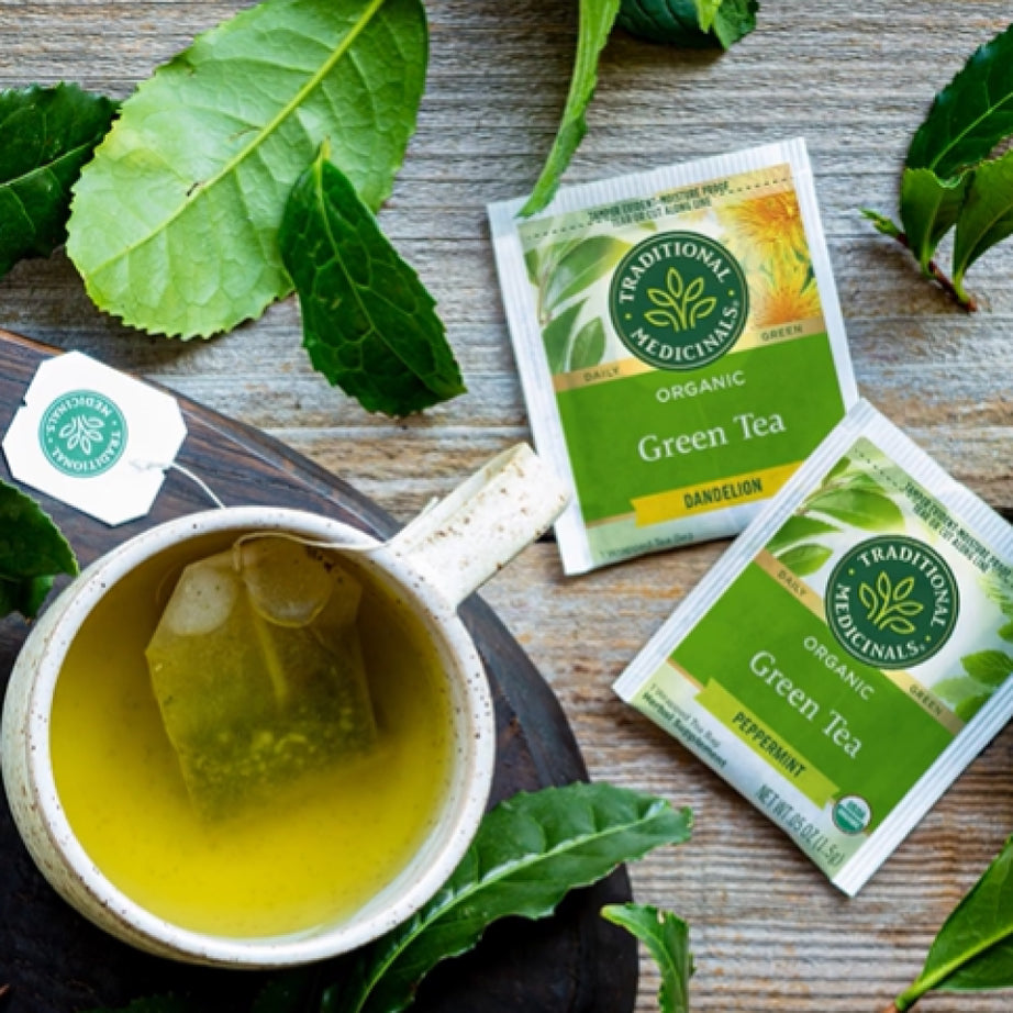 A cup of Green Tea Peppermint with tea bags sits on a wooden surface, surrounded by fresh green leaves.