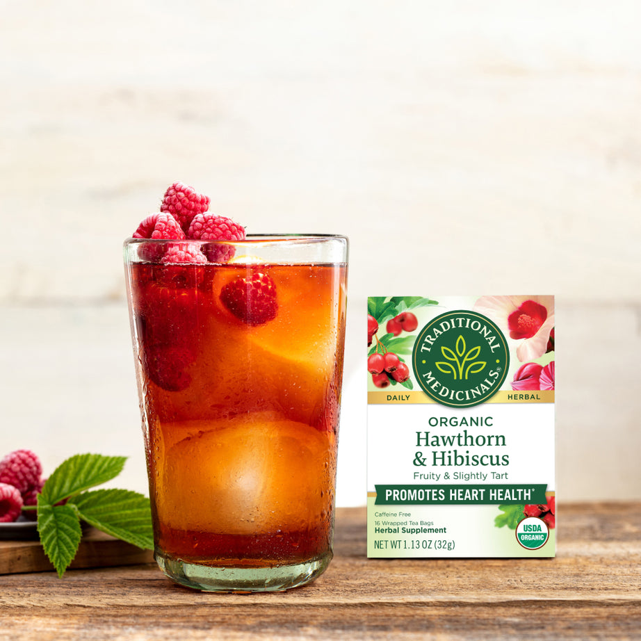 A refreshing glass of iced tea with raspberries sits beside a box of Hawthorn & Hibiscus Tea, combining delightful flavors that support heart health.