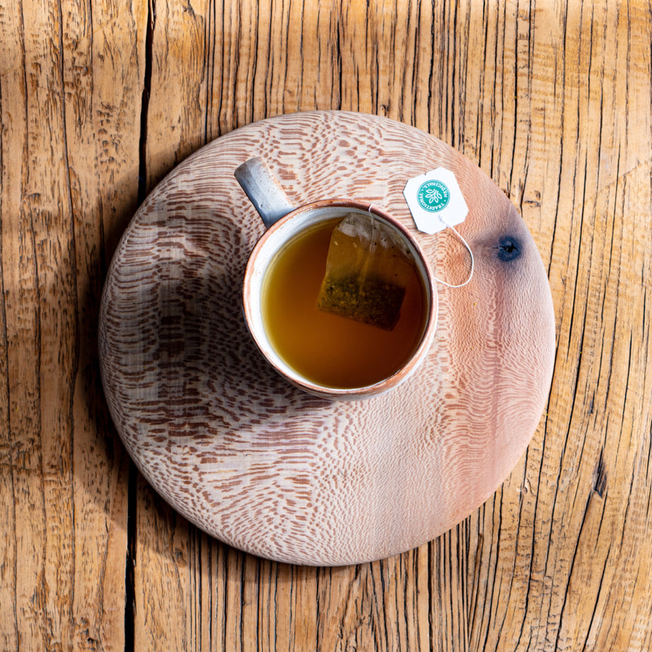 A cup of Herbal Cold Care™ Tea with a tea bag sits on a wooden coaster atop a rustic wooden table, providing soothing comfort.