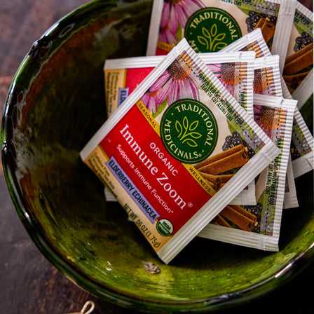 A bowl filled with packets of Immune Zoom® Elderberry Echinacea Tea rests on a wooden surface, providing organic tea benefits. With the essence of elderberry echinacea, it's ideal for immune support.