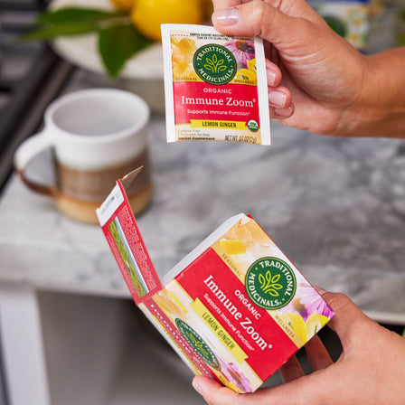 A person holding a packet of Immune Zoom® Lemon Ginger Echinacea Tea over an open box, with a mug and fresh lemon ginger in the background.