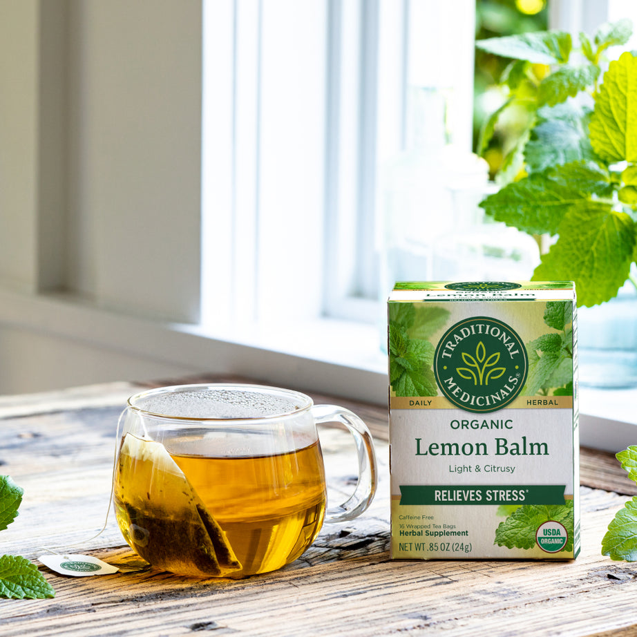 A cup of Lemon Balm Tea, renowned for its soothing effect on the nervous system, rests next to a box of Traditional Medicinals organic lemon balm on a wooden table by the window.