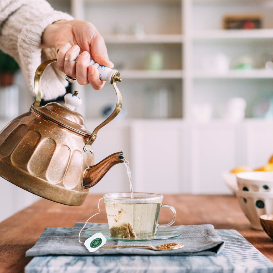 A hand carefully pours steaming hot water from a kettle into a glass cup containing a Moringa with Spearmint & Sage tea bag resting on a wooden table.