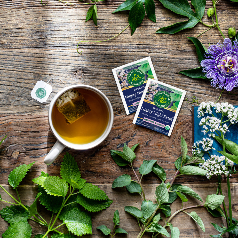 A cup of tea with Nighty Night Extra® Tea bags rests on a wooden table, surrounded by valerian root, fresh herbs, and flowers for a serene deep sleep experience.