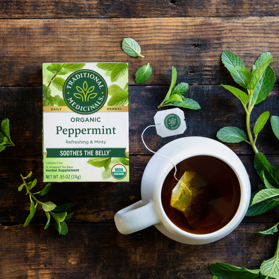 A box of Peppermint Tea sits beside a steaming cup on a wooden table, surrounded by fresh mint leaves. This soothing blend is perfect for easing digestive discomfort while providing a refreshing flavor.