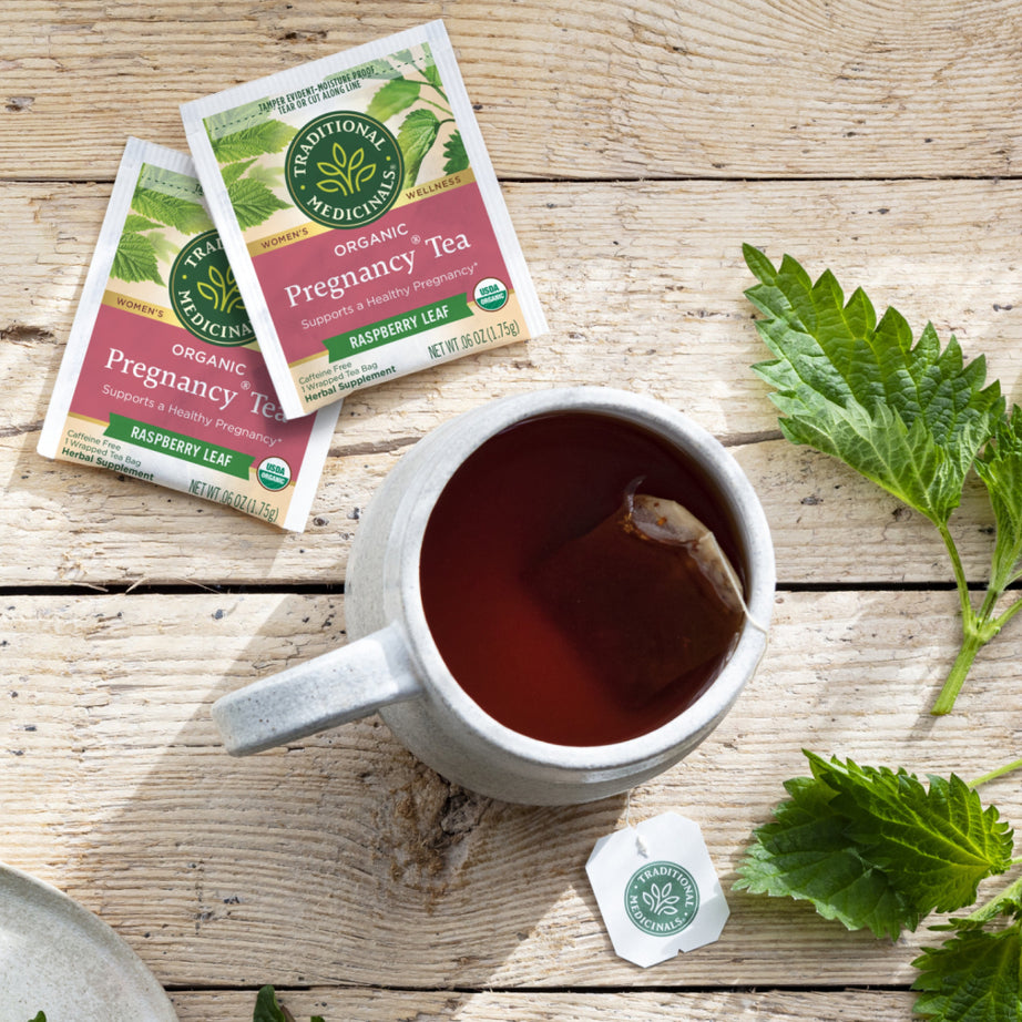 A cup of Pregnancy® Tea rests on a wooden table, infused with the promise of a healthy pregnancy. Surrounded by tea packets and green leaves, this tea offers a soothing moment for soon-to-be mothers.