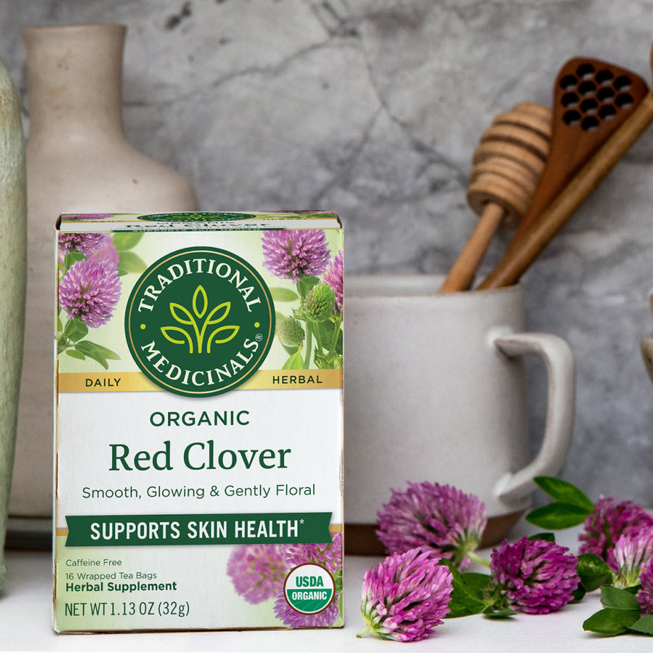 A box of Traditional Medicinals' Red Clover Tea sits surrounded by cups and honey dippers, perfect for enjoying a soothing brew that supports skin health.