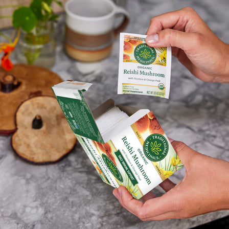 Hands holding a packet of Reishi Mushroom with Rooibos & Orange Peel Tea, renowned as the mushroom of immortality, hover above an open box on a marble countertop.