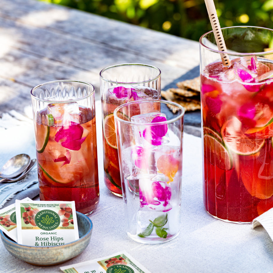 A quartet of glasses filled with refreshing iced Rose Hips & Hibiscus Tea rests on a table outside, beautifully decorated with vibrant flowers and lime slices.