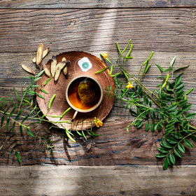 A cup of Smooth Move® Chamomile Tea on a wooden table is surrounded by green leaves and dried roots, offering gentle overnight constipation relief.