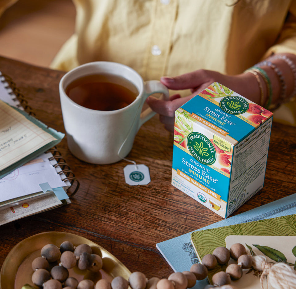 A person enjoys Stress Ease® Immune Tea infused with adaptogens at a table, alongside a box of the same stress-relief tea, notebooks, and decorative beads.