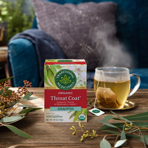 A box of Throat Coat® Eucalyptus Tea sits invitingly on a table, surrounded by eucalyptus leaves and a steaming cup, promoting throat health. A cozy couch in the background beckons for rest and relaxation, offering the perfect setting to enjoy this soothing herbal medicine.