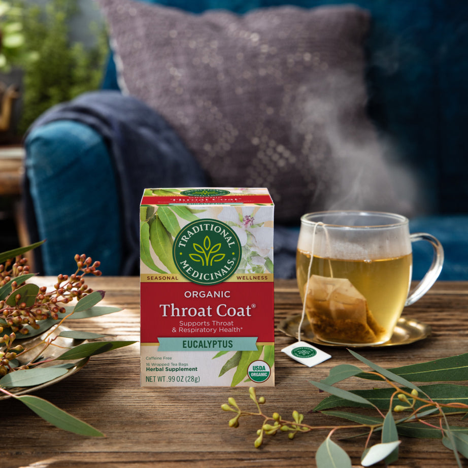 A box of Throat Coat® Eucalyptus Tea sits invitingly on a table, surrounded by eucalyptus leaves and a steaming cup, promoting throat health. A cozy couch in the background beckons for rest and relaxation, offering the perfect setting to enjoy this soothing herbal medicine.