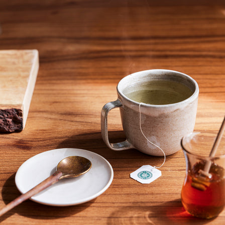 A steaming cup of Tulsi & Ginger Tea, honey jar, and spoon rest on a wooden table, offering the perfect stress support.