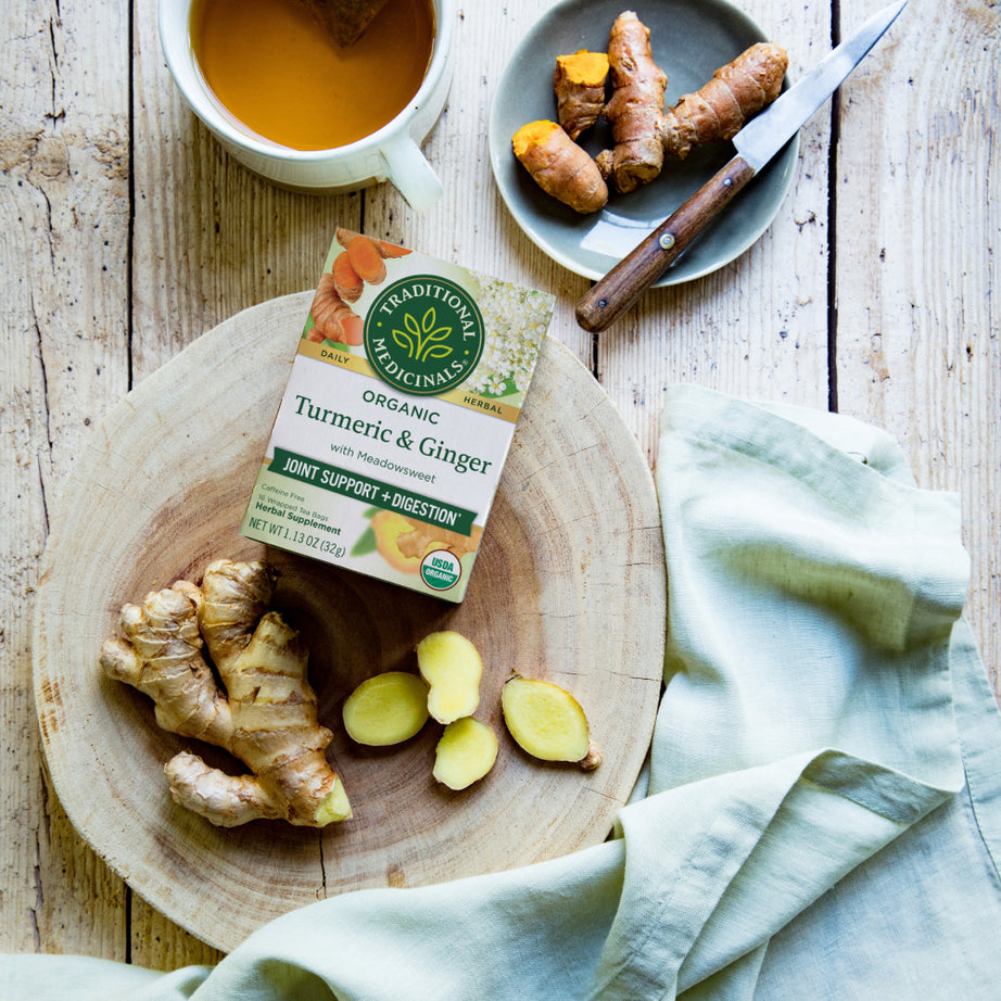 Ginger and turmeric rest on a wooden board next to a cup of meadowsweet tea and a box of Turmeric & Ginger with Meadowsweet Tea on a rustic table.